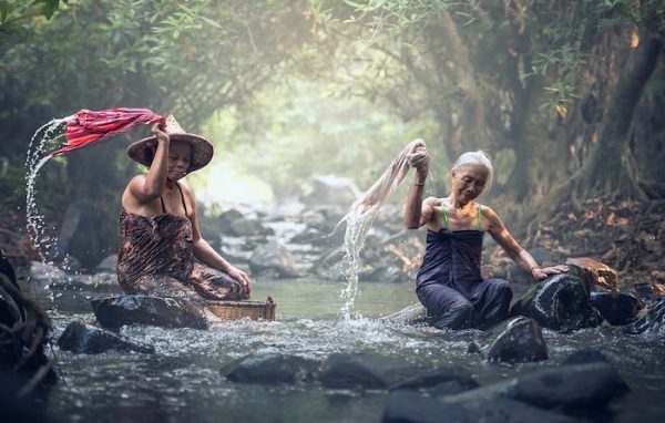 Zwei Frauen schlagen nasse Wäsche am Fluss aus. Zu zweit arbeiten und aufräumen macht mehr Spaß.