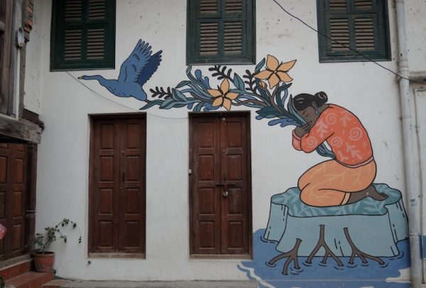 Ein gemaltes Bild an einer Hauswand zeigt eine Frau, die sich an einem Ast mit Blumen festhält. Ein Vogel fliegt davon. Allein zu Haus. 