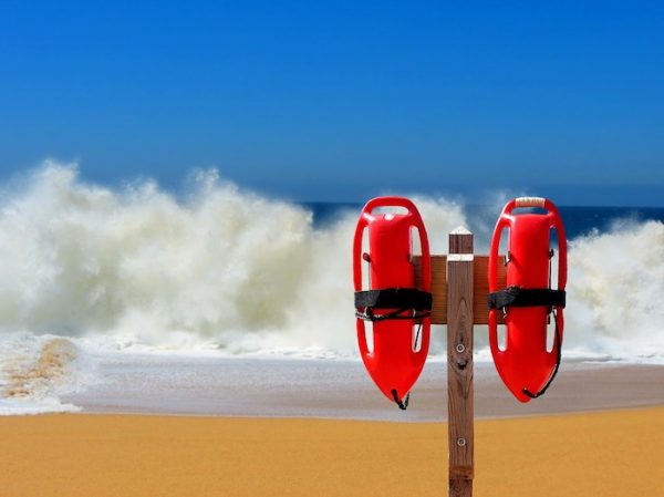 Zwei rote Rettungsbojen hängen an einem Pfahl am Strand, im Hintergrund tosen die Wellen. Hilfe annehmen, auch beim Aufräumen.