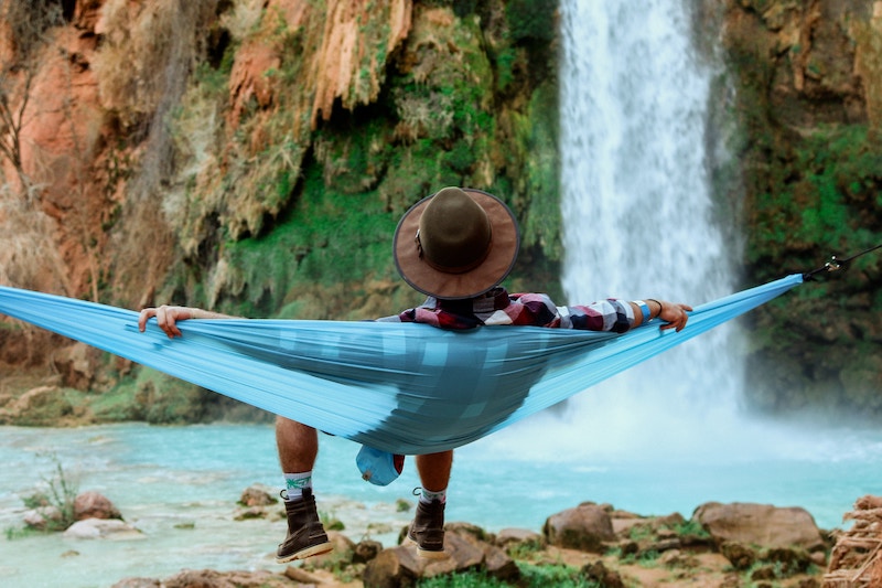 Ein Mann sitzt entspannt in einer blauen Hängematte und betrachtet einen Wasserfall.